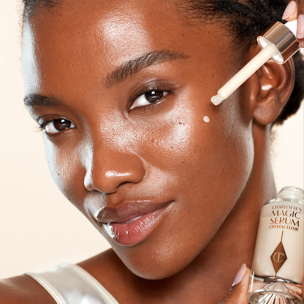 A deep-tone model with brown eyes wearing subtle eye makeup and applying a luminous face serum on her face with a gold-coloured dropper. 